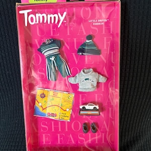 May include: A pink box set of Tommy Little Driver Fashion accessories. The set includes a green and white striped outfit, a gray sweater with a car graphic, a beanie, a road map, a toy car, and black shoes. The box reads "Tommy" and "Fashion Avenue."