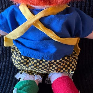 May include: A vintage doll with a bright orange wig, blue shirt, and yellow braces. The doll's trousers are black and yellow chequered, and its socks are green and red. The doll's legs are visible.