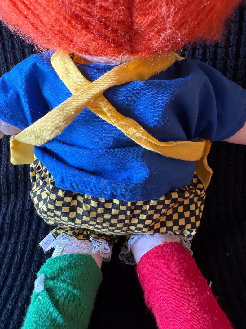 May include: A vintage doll with a bright orange wig, blue shirt, and yellow braces. The doll's trousers are black and yellow chequered, and its socks are green and red. The doll's legs are visible.