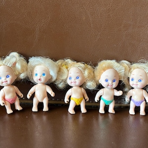 Puede incluir: Cinco pequeñas muñecas de plástico que representan bebés, con pelo rubio y ojos azules. Cada muñeca lleva un pañal de diferente color: rosa, amarillo, turquesa y morado. Las muñecas están dispuestas en fila sobre una superficie marrón.
