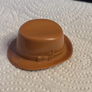 May include: A small, brown fedora hat with a rounded crown and a brim. The hat features a decorative band around the base of the crown. The hat is made of a smooth, solid material.