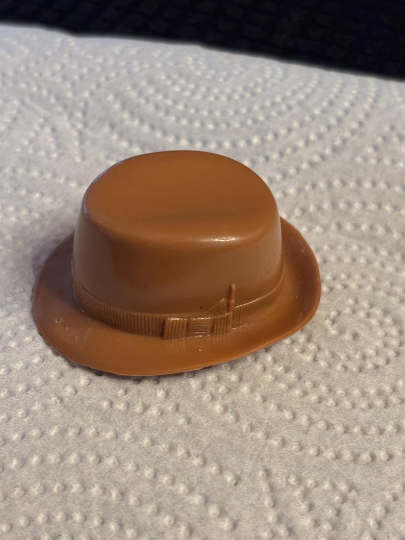 May include: A small, brown fedora hat with a rounded crown and a brim. The hat features a decorative band around the base of the crown. The hat is made of a smooth, solid material.