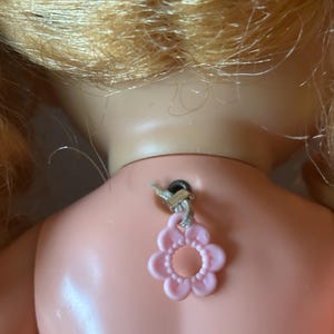 May include: Back of a vintage doll with blonde hair and a pink flower-shaped charm attached to its neck. The doll's skin is a pale pink color, and the charm is suspended by a string.