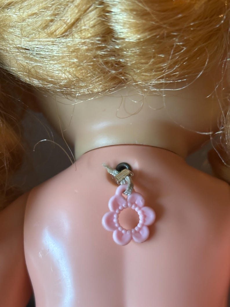 May include: Back of a vintage doll with blonde hair and a pink flower-shaped charm attached to its neck. The doll's skin is a pale pink color, and the charm is suspended by a string.