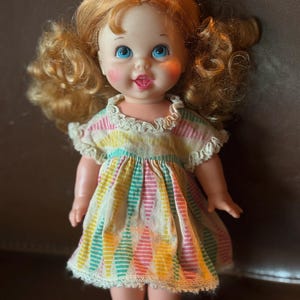May include: A vintage doll with red curly hair and blue eyes. The doll wears a colorful dress with vertical stripes in pink, yellow, and teal. The dress has a white lace trim around the collar and hem. The doll has rosy cheeks and a smiling expression.