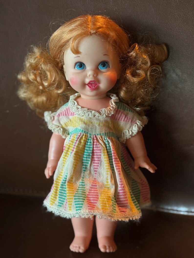 May include: A vintage doll with red curly hair and blue eyes. The doll wears a colorful dress with vertical stripes in pink, yellow, and teal. The dress has a white lace trim around the collar and hem. The doll has rosy cheeks and a smiling expression.