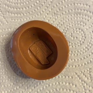 May include: A brown plastic toy hat with a detailed interior. The hat has a circular brim and a central indentation. Inside the hat is a small, rectangular object with a detailed design, possibly a toy component.