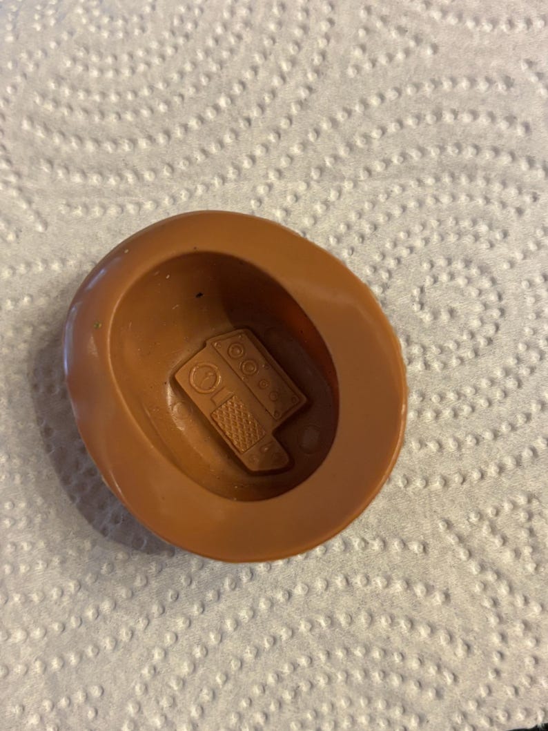 May include: A brown plastic toy hat with a detailed interior. The hat has a circular brim and a central indentation. Inside the hat is a small, rectangular object with a detailed design, possibly a toy component.