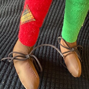 May include: Close-up of a doll's legs and feet. One leg is covered in a red sock with a plaid patch, the other in a green sock. Brown shoes with black laces are tied in bows. The background is a dark, textured fabric.