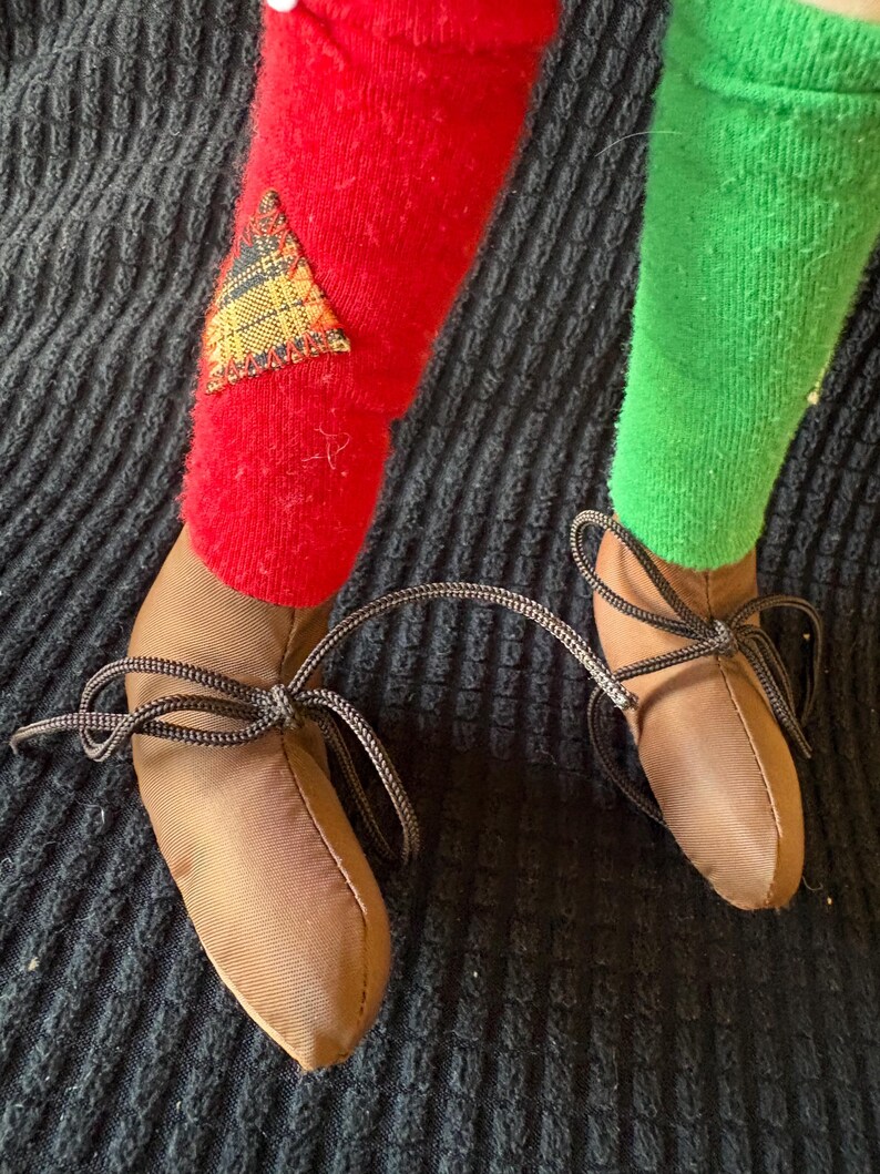 May include: Close-up of a doll's legs and feet. One leg is covered in a red sock with a plaid patch, the other in a green sock. Brown shoes with black laces are tied in bows. The background is a dark, textured fabric.