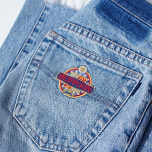 Pode incluir: Um close-up de um bolso traseiro de ganga azul com um remendo bordado em vermelho, amarelo e azul que diz "Special Day", "Buffoon" e "De. 1981".