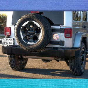 May include: A silver Jeep Wrangler with a spare tire on the back. The spare tire has a black rim and a black and silver tire. The Jeep has a license plate that says "WMNS SPRTS" and a sticker that says "Keep Calm and Wander On".