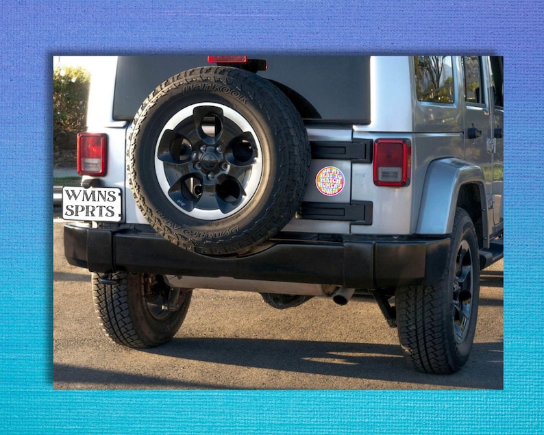 May include: A silver Jeep Wrangler with a spare tire on the back. The spare tire has a black rim and a black and silver tire. The Jeep has a license plate that says "WMNS SPRTS" and a sticker that says "Keep Calm and Wander On".