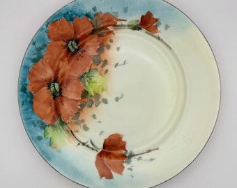 May include: A round ceramic plate with a light blue rim and a cream-colored center. The plate is decorated with hand-painted orange flowers, green leaves, and gray accents. The flowers are the focal point of the design.