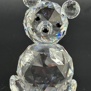 May include: A clear crystal bear figurine with a faceted design. The bear has black eyes and a small black nose. The bear's body, head, and ears are made of multiple crystal facets. The figurine is displayed against a dark background.
