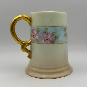 May include: A white porcelain mug with a gold handle and floral design. The mug has a light blue band with pink flowers and green leaves. The base of the mug is a cream color.