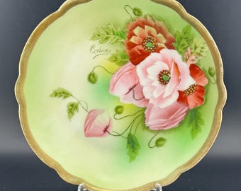 May include: A decorative, scalloped-edge plate with a light green background and a gold rim. The plate features hand-painted pink and red poppies with green leaves. The word "Cochin" is written on the plate.