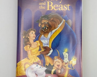 Puede incluir: Una cinta VHS de "La Bella y la Bestia" de Disney. La portada presenta a Bella con un vestido amarillo, la Bestia con un traje azul y otros personajes. El título está en letras doradas sobre un fondo morado.