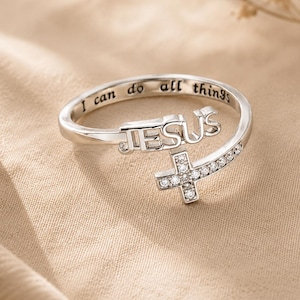 Peut inclure: Une bague ouverte argentée présentant le mot JESUS en lettres en relief et un motif de croix incrusté de petites pierres claires. L'intérieur de l'anneau est gravé avec le texte I can do all things, offrant une finition métallique polie.