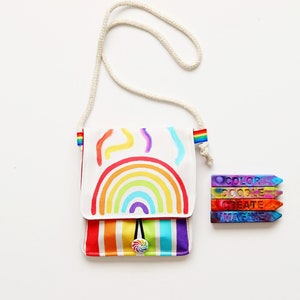 May include: A white crossbody bag with a rainbow design and a matching rainbow strap. The bag has a colorful striped bottom and a lollipop button closure. Next to the bag are four colorful crayons with the words "COLOR DOODLE CREATE IMAGINE".
