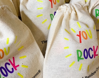 May include: A close-up of several small, white fabric bags with a rainbow-colored "You Rock" design. The bags are stacked on top of each other and have a yellow sunburst design around the text. The text "The Original Rainbow Crayon®" is printed on the bottom of the bag.