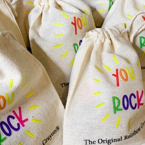 May include: A close-up of several small, white fabric bags with a rainbow-colored "You Rock" design. The bags are stacked on top of each other and have a yellow sunburst design around the text. The text "The Original Rainbow Crayon®" is printed on the bottom of the bag.