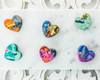 May include: Six heart-shaped crayons in various colors and patterns, including orange, green, blue, and pink, arranged on a white marble surface.