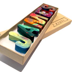 May include: A set of colorful, multi-colored, crayon letters spelling out the name "JAMES" in a brown cardboard box with a label that reads "JAMES".