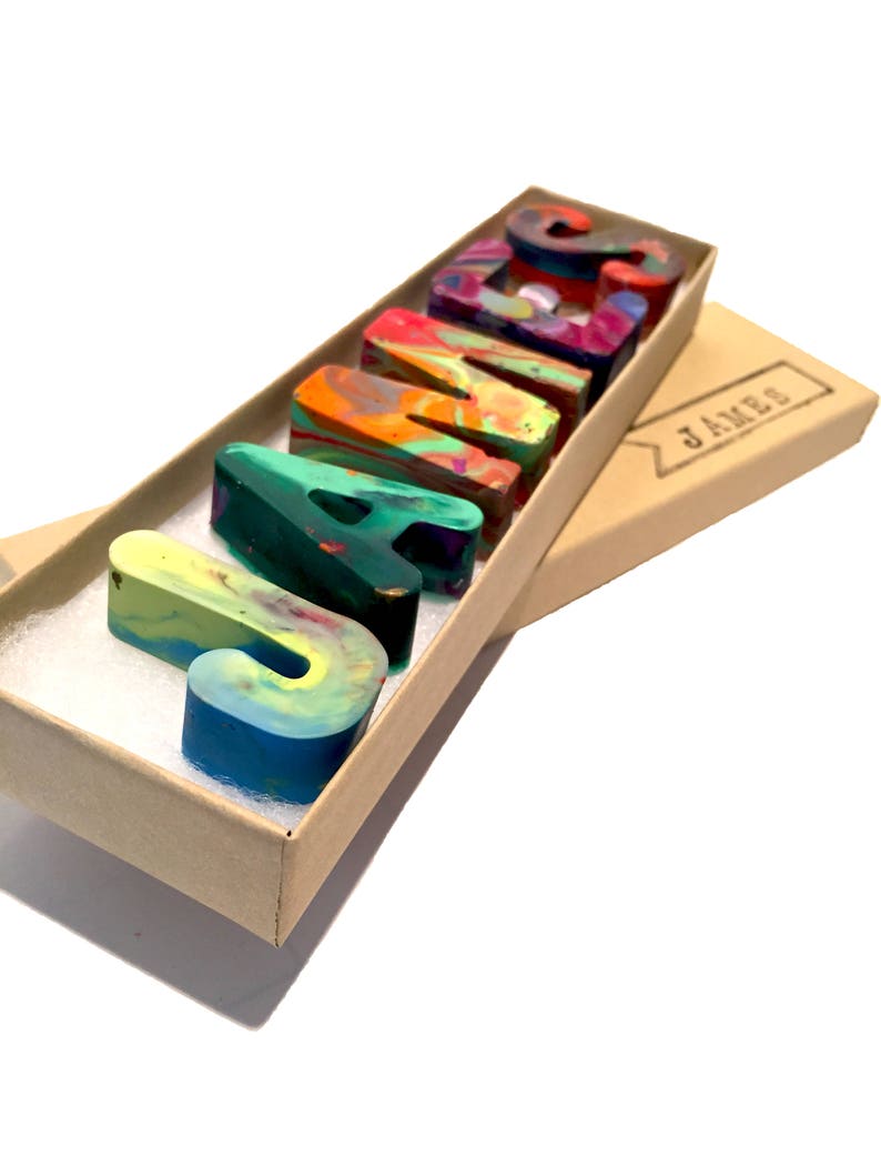 May include: A set of colorful, multi-colored, crayon letters spelling out the name "JAMES" in a brown cardboard box with a label that reads "JAMES".