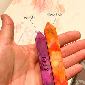 Puede incluir: Dos crayones, uno morado y otro naranja, se sostienen en una mano. El cray&oacute;n morado est&aacute; etiquetado como "mini stix" y el cray&oacute;n naranja est&aacute; etiquetado como "standard stix".