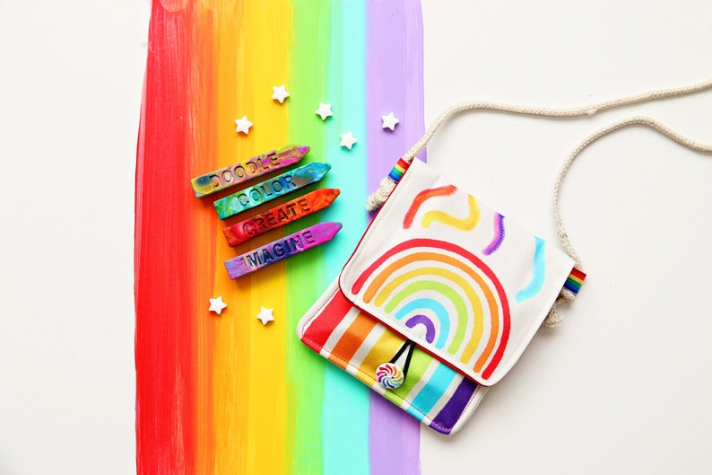 May include: A white crossbody bag with a rainbow design and a rope strap. The bag features a rainbow and colorful accents. Beside the bag are colorful crayons with the words "DOODLE COLOR CREATE IMAGINE" and white star-shaped decorations.