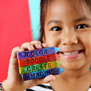 May include: A young person holds a stack of colorful crayons. Each crayon is a different color and has a word carved into it: COLOR, DOODLE, CREATE, and IMAGINE. The background is red and blue.