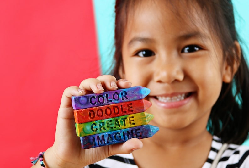 May include: A young person holds a stack of colorful crayons. Each crayon is a different color and has a word carved into it: COLOR, DOODLE, CREATE, and IMAGINE. The background is red and blue.