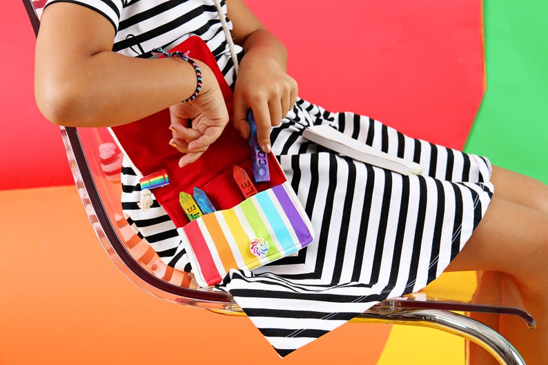 May include: A colorful crayon bag with a rainbow striped pocket, red interior, and a white strap. The bag contains several crayons in various colors. The person is wearing a black and white striped dress and a bracelet.