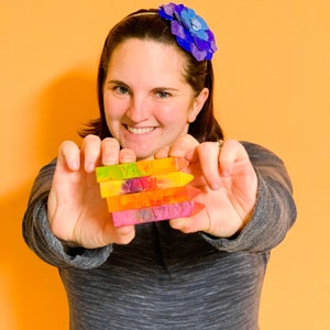 Puede incluir: Una persona sostiene una pila de cinco crayones de colores, cada uno con la palabra "YAY" escrita en &eacute;l. Los crayones est&aacute;n dispuestos en un patr&oacute;n de arco&iacute;ris, con el cray&oacute;n superior amarillo y el cray&oacute;n inferior rosa.