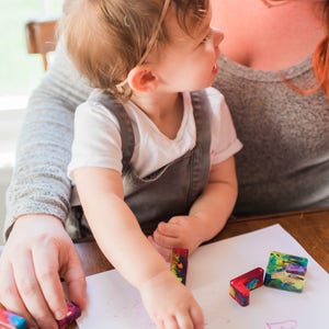 May include: A young child sits on a person's lap and draws on a white sheet of paper with colorful crayons. The child is wearing a grey overall and a pink headband. The word "friend" is written on the paper.