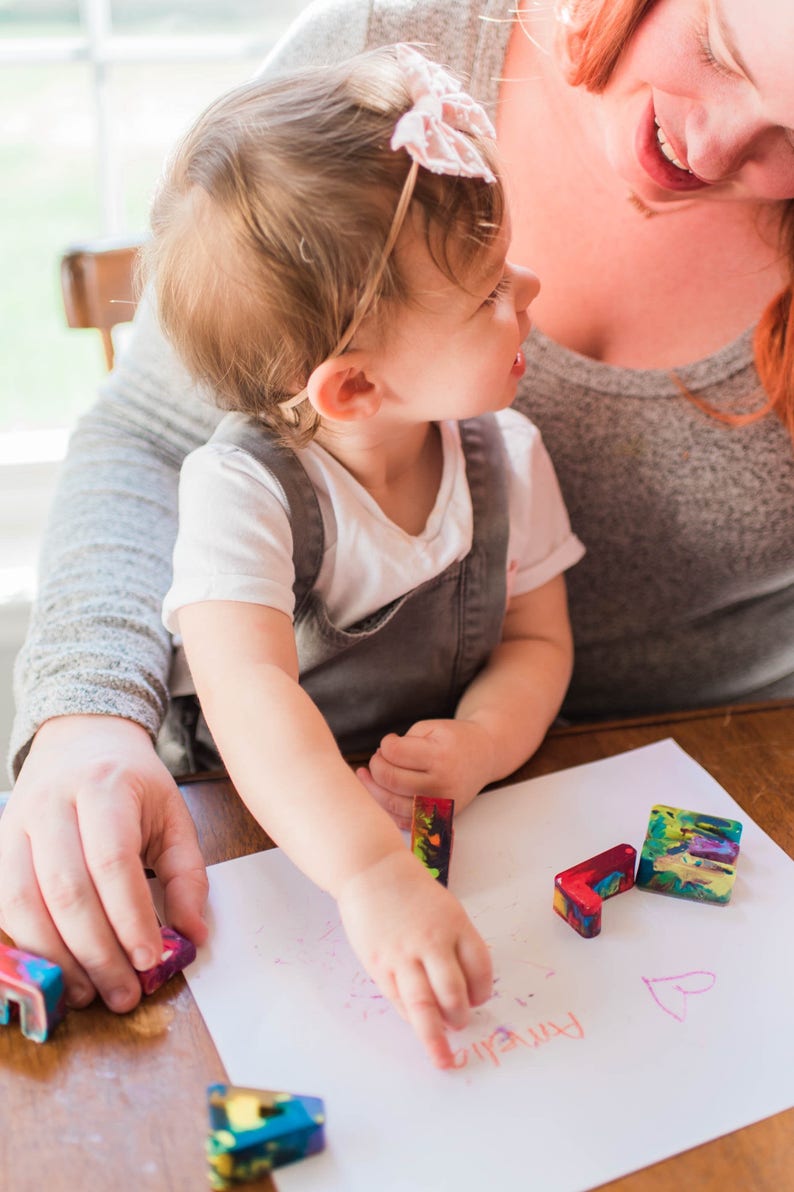 May include: A young child sits on a person's lap and draws on a white sheet of paper with colorful crayons. The child is wearing a grey overall and a pink headband. The word "friend" is written on the paper.