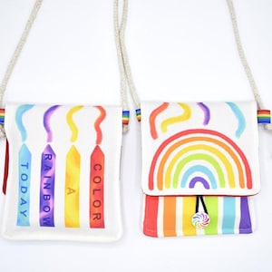 May include: Two white canvas crossbody bags with rainbow designs. One bag features the text "TODAY A RAINBOW COLOR" on crayon-shaped graphics. The other bag has a rainbow and colorful stripes. Both have rope straps and rainbow accents.