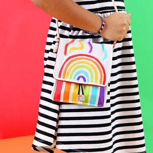 May include: A white crossbody bag with a rainbow design. The bag has a flap with a rainbow and colorful stripes. The strap is a cream-colored rope. The bag is held by a person wearing a black and white striped dress.