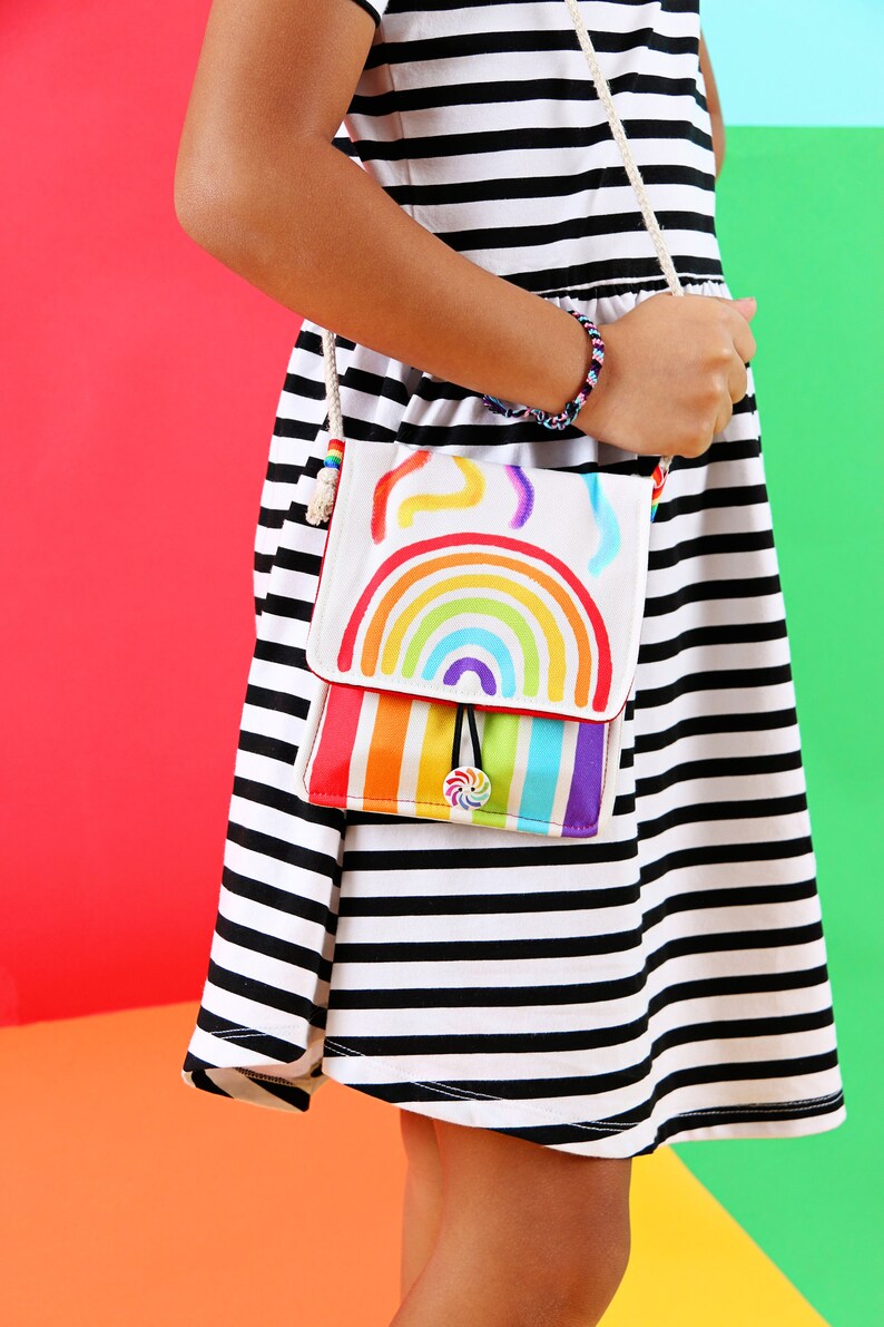 May include: A white crossbody bag with a rainbow design. The bag has a flap with a rainbow and colorful stripes. The strap is a cream-colored rope. The bag is held by a person wearing a black and white striped dress.