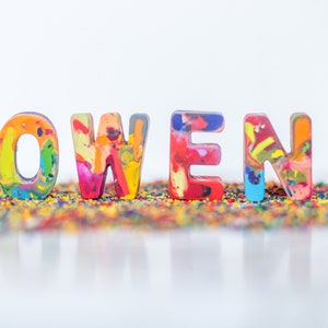May include: The name "OWEN" spelled out in colorful, multi-colored crayon letters on a bed of rainbow-colored crayon shavings.