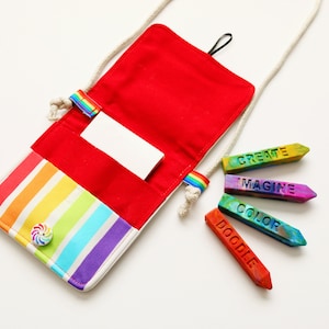May include: A red and rainbow striped crossbody bag with a white cord strap. The bag is open, revealing a red interior and a white card. Colorful crayons with the words "CREATE", "IMAGINE", "COLOR", and "DOODLE" are next to the bag.