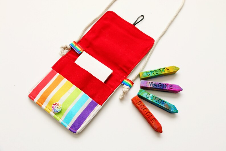 May include: A red and rainbow striped crossbody bag with a white cord strap. The bag is open, revealing a red interior and a white card. Colorful crayons with the words "CREATE", "IMAGINE", "COLOR", and "DOODLE" are next to the bag.