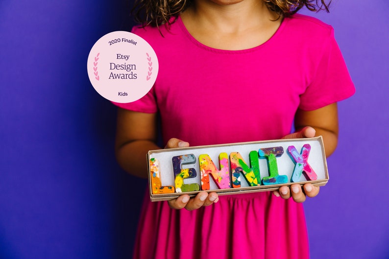 May include: A child holds a box with the name "LENNIX" spelled out in colorful crayon letters. The box is made of cardboard and has a brown finish. The child is wearing a pink shirt and is standing in front of a purple background. The box has a 2020 Etsy Design Awards Finalist sticker on it.