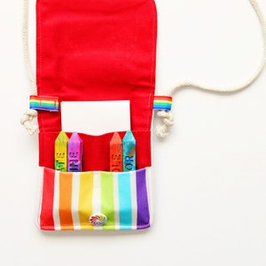 May include: A red canvas crossbody bag with a rainbow striped pocket and a white rope strap. The bag contains four colorful crayons with the words "ART" and "COLOR" printed on them. The bag has a rainbow accent on each side.