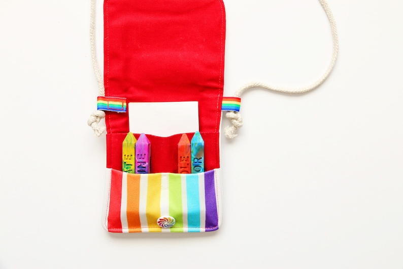 May include: A red canvas crossbody bag with a rainbow striped pocket and a white rope strap. The bag contains four colorful crayons with the words "ART" and "COLOR" printed on them. The bag has a rainbow accent on each side.