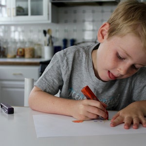 May include: A young boy sits at a table, drawing on a white sheet of paper with a red crayon. He is wearing a grey t-shirt with the word "Ocean" printed on it. Two small, colorful rectangular objects are on the table in front of him.