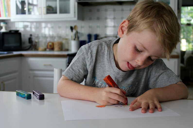 May include: A young boy sits at a table, drawing on a white sheet of paper with a red crayon. He is wearing a grey t-shirt with the word "Ocean" printed on it. Two small, colorful rectangular objects are on the table in front of him.