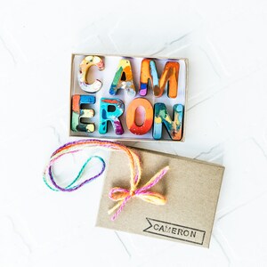 May include: A small box with a colorful set of crayon letters spelling out the name "CAMERON". The box is tied with a multi-colored string and has the name "CAMERON" printed on the front.