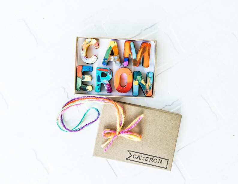 May include: A small box with a colorful set of crayon letters spelling out the name "CAMERON". The box is tied with a multi-colored string and has the name "CAMERON" printed on the front.